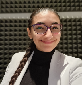 Virginia Chachati wearing glasses with a long brown plait and a black polo neck with a white suit jacket and sound proof background